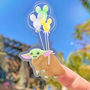 May include: A clear sticker of a green baby character holding onto three Mickey Mouse-shaped balloons. The balloons are green, white, and yellow.