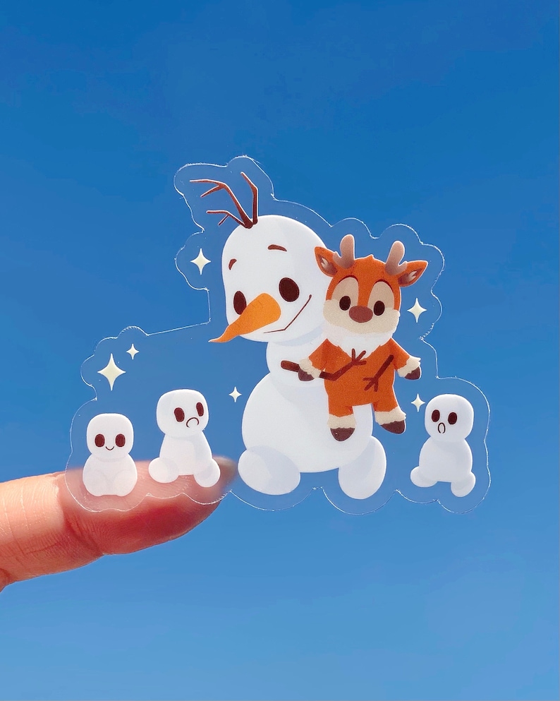 Puede incluir: Una pegatina transparente con un mu&ntilde;eco de nieve con un reno montando en su espalda. Hay tres mu&ntilde;ecos de nieve m&aacute;s peque&ntilde;os en el fondo. La pegatina est&aacute; sobre un fondo azul.