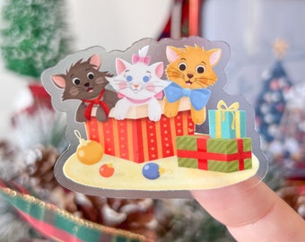 May include: A clear sticker featuring three cartoon cats in a red and gold striped gift box. The cats are brown, white, and orange, with bows and ribbons. Christmas ornaments and wrapped gifts are also visible.