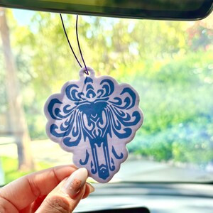 May include: A purple and blue car air freshener shaped like a haunted mansion with a ghost inside. The air freshener is hanging from the rearview mirror of a car.
