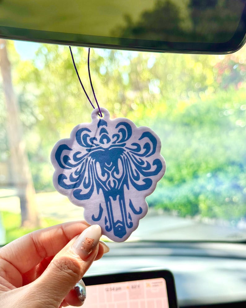May include: A purple and blue car air freshener shaped like a haunted mansion with a ghost inside. The air freshener is hanging from the rearview mirror of a car.