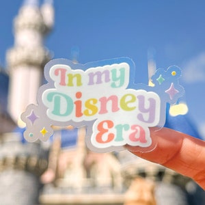 May include: A clear sticker with the text "In my Disney Era" in rainbow colors. The sticker is held in front of a blurred image of Cinderella's Castle.