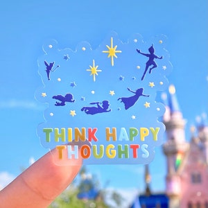 Op de afbeelding: Een transparante sticker met een blauw, geel en wit ontwerp met silhouetten van personages uit de film Peter Pan. De sticker heeft de tekst "Think Happy Thoughts" in regenboogkleuren.