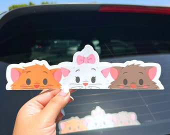 May include: Three cartoon cat faces peeking over the top of a car window. The cat on the left is orange, the cat in the middle is white with a pink bow, and the cat on the right is brown.
