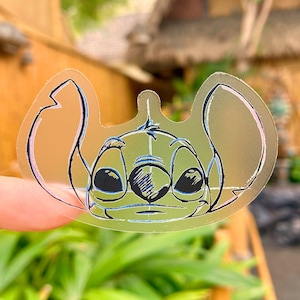 May include: A clear sticker with a blue and black outline of Stitch, a character from the Disney movie Lilo & Stitch. The sticker is on a person's finger.