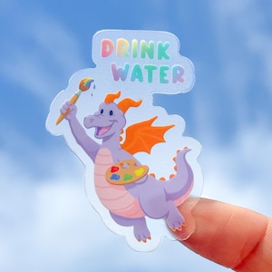 May include: A clear sticker featuring a cartoon dragon with a paintbrush and a palette. The dragon is purple with orange wings and horns, holding a paintbrush with rainbow paint. The sticker has the words "DRINK WATER" in colorful letters.