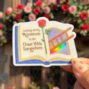 May include: A sticker in the shape of an open book with the text "Leaving on an Adventure to the Great Wide Somewhere." A red rose and a ladder are featured. The background is a window with a rainbow effect.
