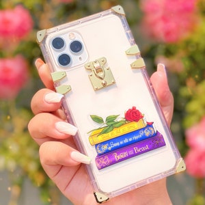 May include: Clear phone case with a gold trim and a gold lock. The case has a sticker of a stack of books with a red rose on top. The books have the titles "Tale as old as time", "Song as old as rhyme", and "Beauty and the Beast".