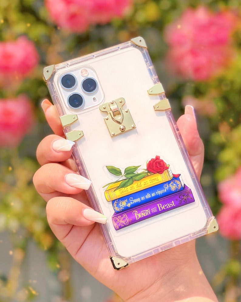 May include: Clear phone case with a gold trim and a gold lock. The case has a sticker of a stack of books with a red rose on top. The books have the titles "Tale as old as time", "Song as old as rhyme", and "Beauty and the Beast".