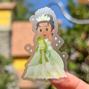 May include: A sticker featuring a cartoon illustration of a princess in a green and yellow gown, holding a green frog. The princess has dark hair and a crown. The sticker has a silver outline with star accents.