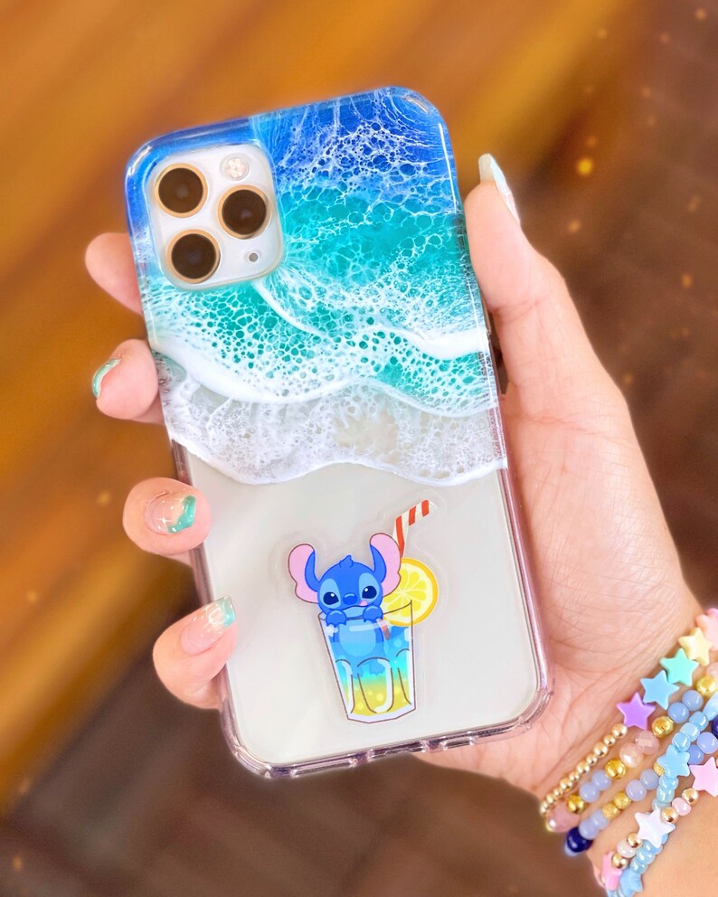 Puede incluir: Una funda transparente para tel&eacute;fono con un dise&ntilde;o de ola de oc&eacute;ano azul y verde. Un personaje de dibujos animados azul sostiene un vaso de limonada con una rodaja de lim&oacute;n y una pajita roja. El personaje est&aacute; en una pegatina transparente en la parte posterior de la funda.