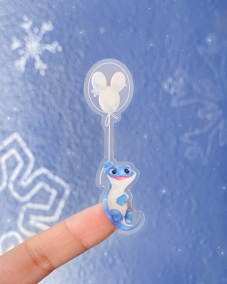 Pu&ograve; includere: Un adesivo trasparente con una salamandra blu che tiene un palloncino bianco con una silhouette di Topolino. L'adesivo &egrave; su un dito su uno sfondo blu.