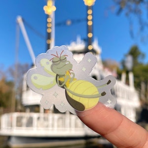 Puede incluir: Una pegatina transparente con una ilustración de dibujos animados de una luciérnaga verde con alas amarillas y un cuerpo brillante. La luciérnaga se representa con los ojos cerrados y una sonrisa en la cara. La pegatina se coloca en un dedo frente a un fondo borroso de un barco blanco.