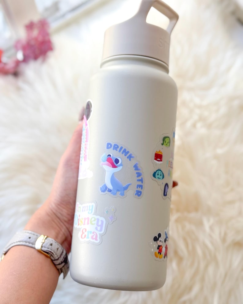 May include: A light grey reusable water bottle with a black lid and handle. The bottle is covered in colourful stickers, including a blue and white character, a Mickey Mouse sticker, and a sticker that says "Drink Water".