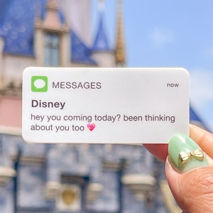 May include: A white sticker with a text message bubble design. The message reads "Disney hey you coming today? been thinking about you too ❤️". The sticker is held in front of a blurred background of a castle.