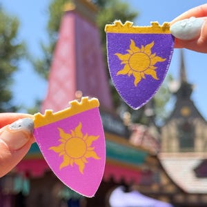 Puede incluir: Dos pequeños parches de tela en forma de escudo. Uno es morado con un diseño de sol dorado, el otro es rosa con un diseño de sol dorado. Ambos tienen un borde superior festoneado dorado. Los parches se sostienen contra un fondo borroso.