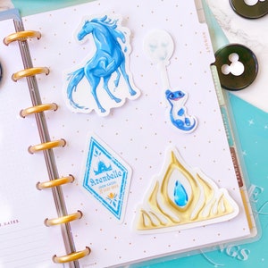 Pu&ograve; includere: Un set di quattro adesivi a tema Frozen che includono un cavallo blu, una salamandra blu, uno stemma blu e bianco con il testo "Arendelle Open Gates 22.12.2013" e una corona dorata con un gioiello blu.