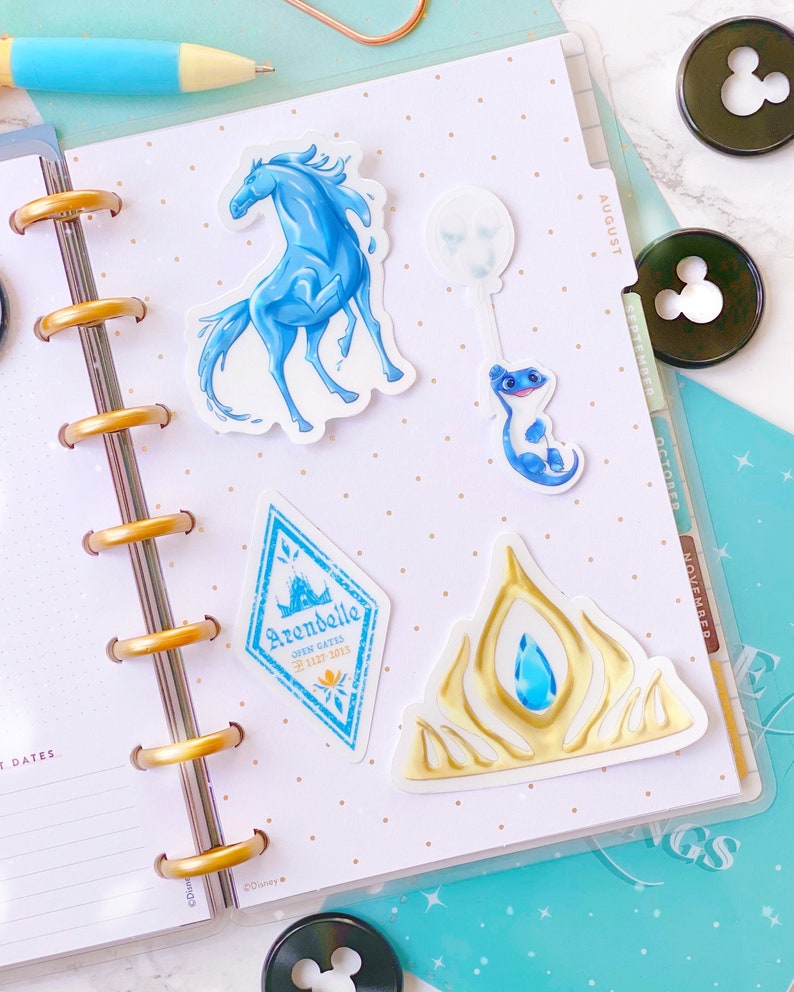 Pu&ograve; includere: Un set di quattro adesivi a tema Frozen che includono un cavallo blu, una salamandra blu, uno stemma blu e bianco con il testo "Arendelle Open Gates 22.12.2013" e una corona dorata con un gioiello blu.