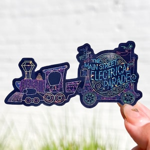 以下が含まれることがあります： 黒いビニールステッカーで、キラキラと虹色の電車のデザインと、円の中に「The Main Street Electrical Parade」という文字が書かれています。