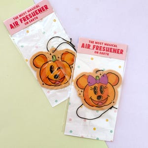 May include: Two hanging air fresheners in clear plastic packaging with pink header cards reading THE MOST MAGICAL AIR FRESHENER ON EARTH. One features an orange pumpkin with mouse ears, the other a pumpkin with mouse ears and a purple bow.