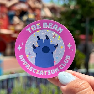 May include: A round sticker with the text "TOE BEAN APPRECIATION CLUB" in white on a pink border. The center features a blue paw print on a glittery background. Two pink stars are on either side of the text.