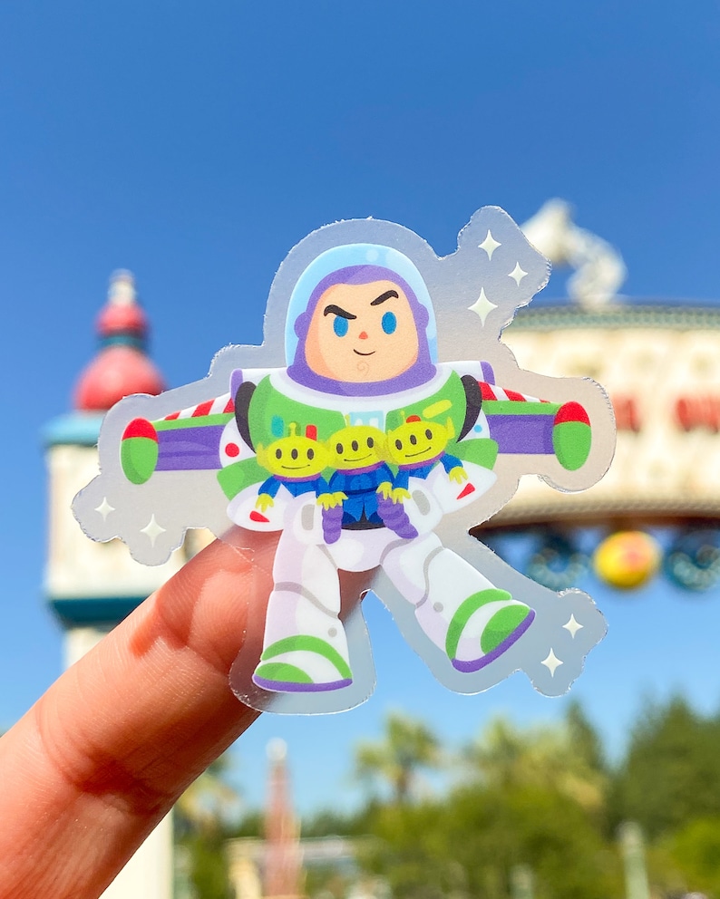 May include: A clear sticker featuring a cartoon illustration of Buzz Lightyear from Toy Story. Buzz is wearing his space ranger suit and holding three green aliens. The sticker has a sparkly, holographic effect.
