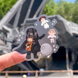 May include: A clear sticker featuring a cartoon Darth Vader holding a small cartoon Obi-Wan Kenobi. The sticker also includes a Death Star, Princess Leia, and Luke Skywalker.