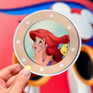 May include: A round, tan-colored sticker featuring a cartoon illustration of Ariel from The Little Mermaid, looking up with a dreamy expression. Flounder is peeking from behind. The sticker is held by a hand with a yellow nail.