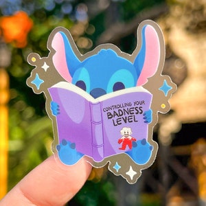 Puede incluir: Un personaje azul de Stitch, de Lilo & Stitch, está leyendo un libro morado que dice "Controlando tu nivel de maldad". El libro tiene una pequeña ilustración de un personaje rojo con una cara blanca.