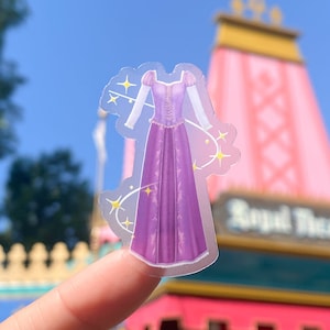 May include: A clear sticker with a purple dress with a white bodice and long sleeves. The dress has a sparkly design and is surrounded by yellow stars. The sticker is on a finger in front of a pink and yellow building.