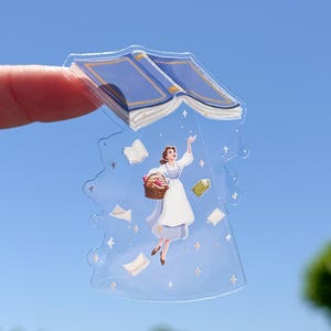 May include: A clear sticker featuring a cartoon illustration of a woman in a white dress reaching towards an open blue book. The sticker has a basket, floating books, and star accents, set against a blue sky background.