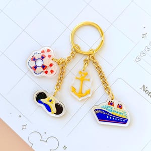 May include: A gold-toned metal keychain featuring four acrylic charms attached by small chains. The charms include a red and white life preserver, a yellow anchor, a blue and yellow sailor hat, and a blue cruise ship with red smokestacks.