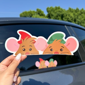 May include: A sticker featuring two cartoon mice peeking out from behind a car window. The mouse on the left is wearing a red hat and the mouse on the right is wearing a green hat.