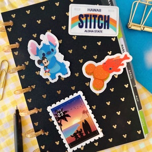 May include: A black planner with gold Mickey Mouse ears printed on the cover. The planner has a clear plastic cover with tabs for each month of the year. There are three stickers on the planner: a blue Stitch character sticker, a red and orange fish sticker, and a vintage-style postcard sticker with a sunset scene.