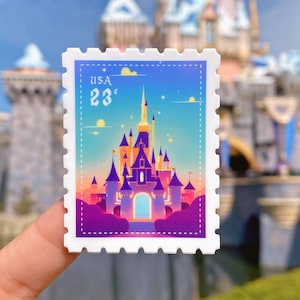 以下が含まれることがあります： 青、紫、ピンクのグラデーションの背景を持つ白いステッカー。ステッカーには、シンデレラ城の漫画イラストが描かれており、左上に「USA 23¢」の文字があります。