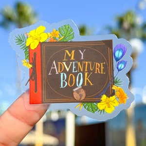 Puede incluir: Una pegatina transparente con una ilustración de libro marrón. El libro tiene el texto "MY ADVENTURE BOOK" en la portada. La pegatina está rodeada de flores y hojas de colores.
