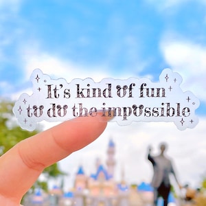 Puede incluir: Una pegatina transparente con el texto "It's kind of fun to do the impossible" en letras negras. La pegatina tiene un borde brillante y se sostiene frente a un fondo borroso.