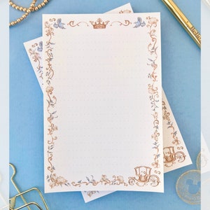 May include: Two sheets of lined stationery with a floral and crown design. The border features brown and blue flowers, butterflies, and a carriage illustration. The stationery is on a light blue surface with a gold pen and jewelry.