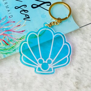 May include: A blue and white seashell-shaped keychain with a gold chain and a Mickey Mouse silhouette inside. The keychain is made of clear acrylic and has a shimmery finish.