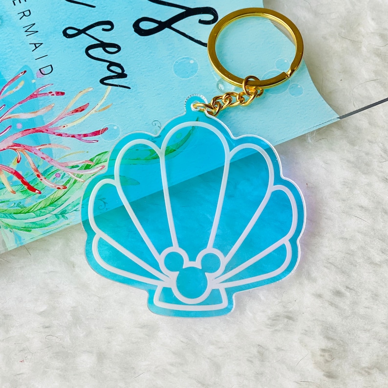 May include: A blue and white seashell-shaped keychain with a gold chain and a Mickey Mouse silhouette inside. The keychain is made of clear acrylic and has a shimmery finish.