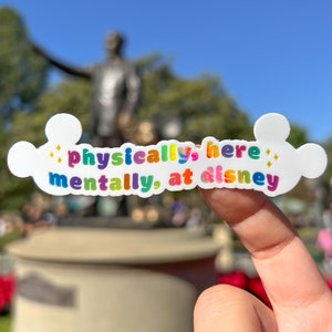 以下が含まれることがあります： 虹色の文字で「physically here, mentally at disney」と書かれた白いステッカー。文字の両側にキラキラが輝いています。ステッカーは、像と木々のぼやけた背景の前で、指で持ち上げられています。