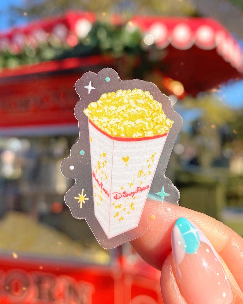 K&ouml;nnte beinhalten: Ein transparenter Aufkleber mit einem wei&szlig;en und roten Popcorn-Eimer, gef&uuml;llt mit gelbem Popcorn. Der Eimer tr&auml;gt den Schriftzug "Disney Parks" und ist von goldenen Sternen umgeben.