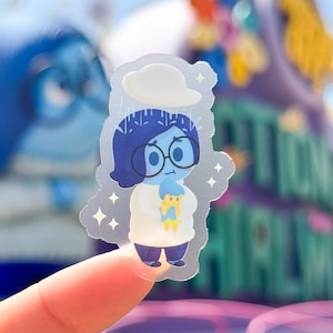 May include: A clear sticker featuring a cartoon character with blue hair, wearing glasses and a white shirt, holding a blue and yellow cartoon character. The character is surrounded by a light blue background with white sparkles.