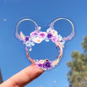 以下が含まれることがあります： 透明なステッカーに、紫、ピンク、ブルーの色の花輪が描かれています。花輪はミッキーマウスの耳の形をしていて、金色のアクセントが付けられています。