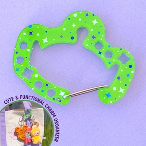 May include: A bright green charm organizer with a heart-shaped design, adorned with blue and white star and dot patterns. The text "CUTE & FUNCTIONAL CHARM ORGANIZER" is visible. A silver-colored metal clasp is attached.