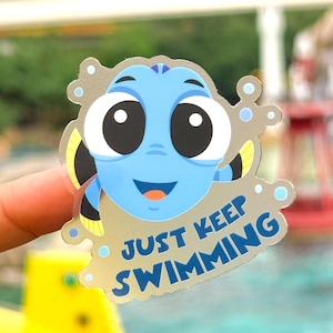May include: A sticker featuring a cartoon blue fish with large black eyes and the text "JUST KEEP SWIMMING" in blue. The fish has yellow and black fins and a smiling orange mouth. The sticker has a silver border.