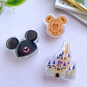 May include: Three clear acrylic Disney-themed charms. One is a black Mickey Mouse head with a red circle, another is a Mickey Mouse waffle, and the last is a castle with pink, blue, and gold accents. The charms are on a light purple surface.