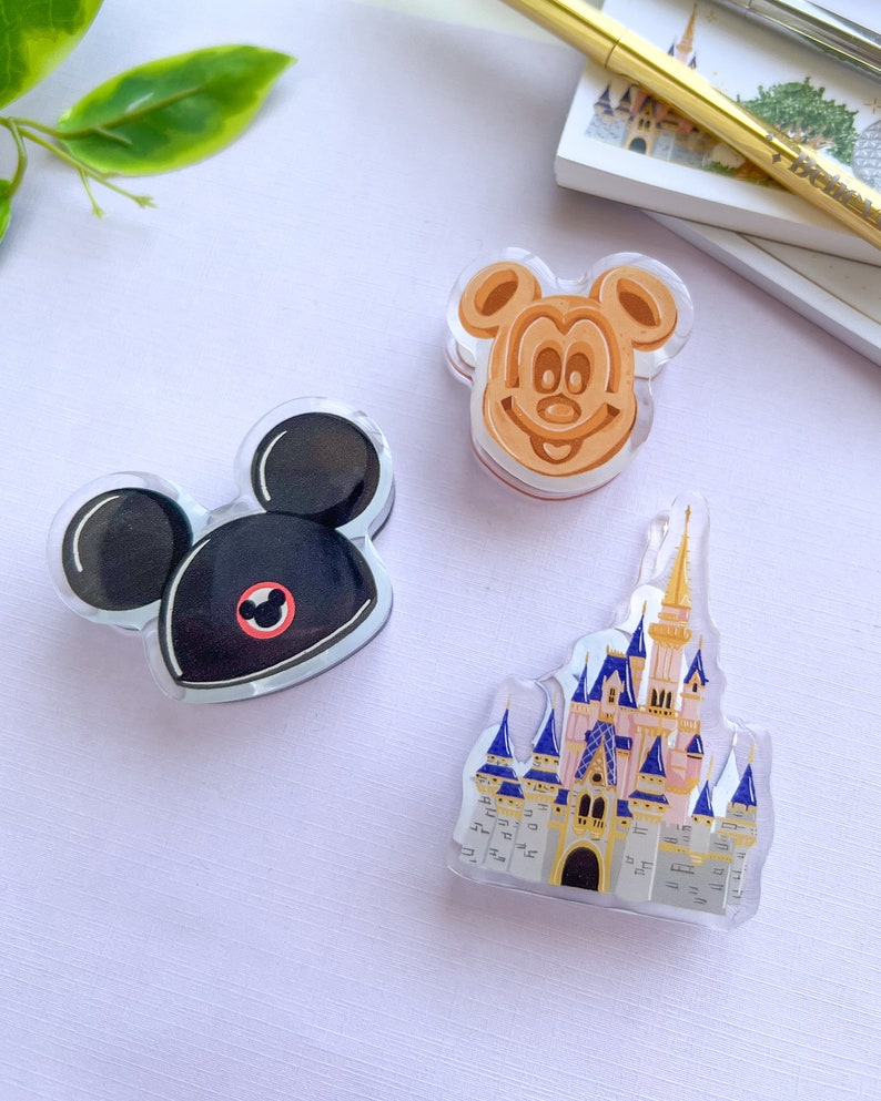 May include: Three clear acrylic Disney-themed charms. One is a black Mickey Mouse head with a red circle, another is a Mickey Mouse waffle, and the last is a castle with pink, blue, and gold accents. The charms are on a light purple surface.