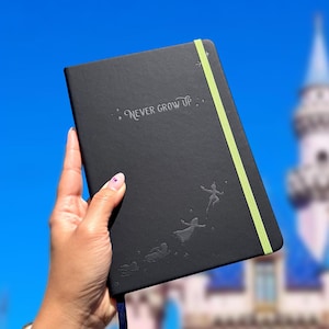 May include: Black journal with the words "Never Grow Up" and embossed Peter Pan-themed silhouettes. The journal has a light green elastic band closure and a blue ribbon bookmark.