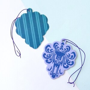 May include: Two car air fresheners, one with a blue and white striped pattern and the other with a purple background and a blue silhouette of a ghost.